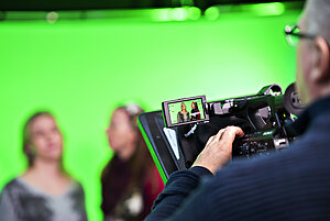 two persons in front of a green screen being filmed by another person