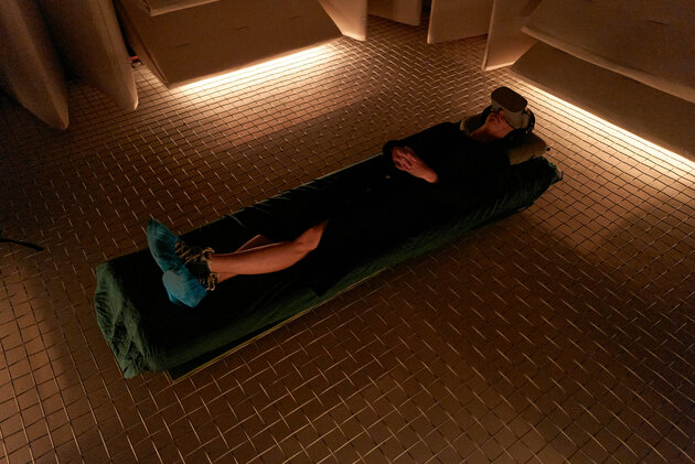 woman lying down on a matress on a mesh floor, wearing a VR headset. Soft light coming from below the walls made of some kind of sound insulation.