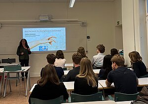 classroom setting. A woman standing in front of a projected image on witheboard. You see students backs, looking at the whiteboard.
