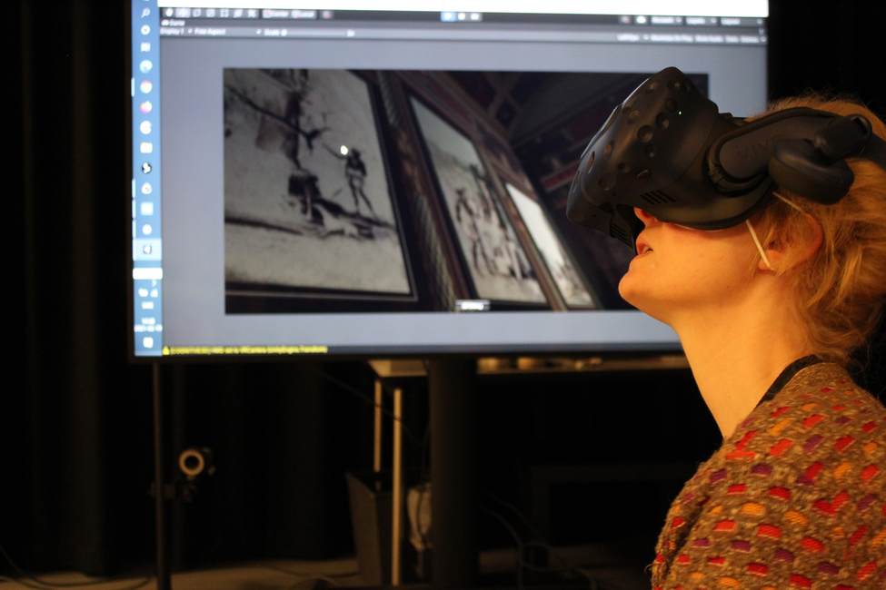 person seen in profile from the left, looking slightly up and wearing VR-headset, in the background a large screen 